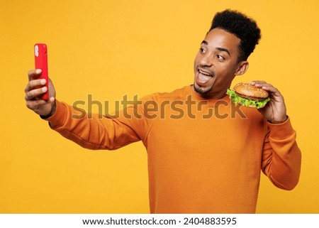Young man wear orange sweatshirt casual clothes eat burger do selfie shot mobile cell phone open mouth isolated on plain yellow background. Proper nutrition healthy fast food unhealthy choice concept