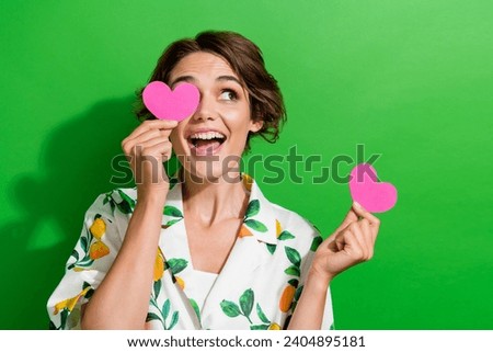Photo of adorable excited girl dressed print shirt holding two pink hearts cover eye looking empty space isolated green color background