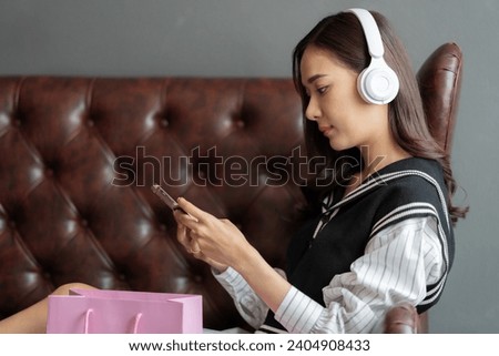 Close-up photo of young Asian woman with long hair being an online seller Wearing headphones and using a credit card to place an order with a smartphone Sitting on the sofa on vacation