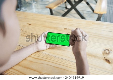                         Man's hand is holding a smartphone on a wooden table with a green screen       