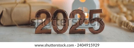 Happy new year 2025 postcard. Golden digits in front of Christmas lights, candles, gift box. White wooden background. Copy space for greeting text banner