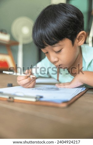 Close up portrait of Asian boy with black bangs, black eyes with a smiling face wearing a light green and lying on the floor of his house drawing a picture of a girl with braids. Education concept.