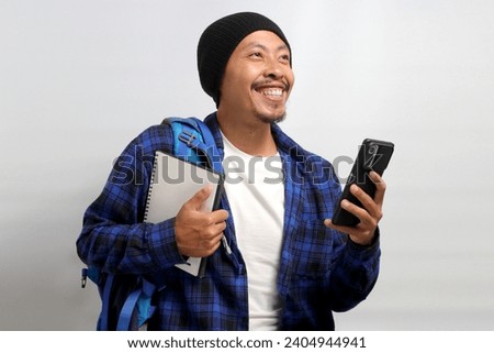 Cheerful Asian student, dressed in a beanie hat and casual shirt and carrying a backpack, holds a notebook and a mobile phone, looking towards an empty space with a contemplative expression