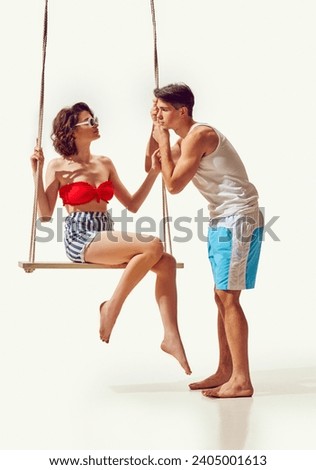 Young muscular man tenderly looking at beautiful young woman sitting on swing against white background. Love and friendship. Concept of summer vacation, travelling, retro style, fashion