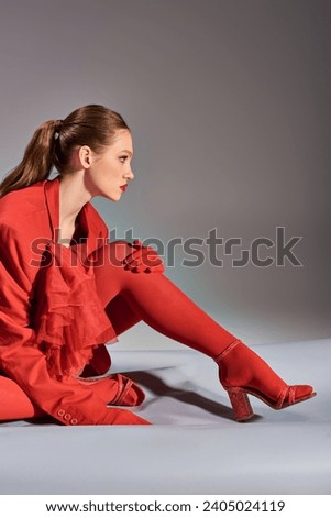 side view of stylish young model in red outfit with tights and high heels sitting on grey background Royalty-Free Stock Photo #2405024119