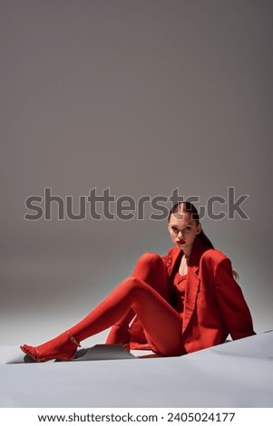 full length of stylish young woman in red attire with gloves and blazer posing on grey background Royalty-Free Stock Photo #2405024177