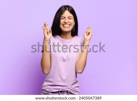 young pretty hispanic woman smiling and anxiously crossing both fingers, feeling worried and wishing or hoping for good luck