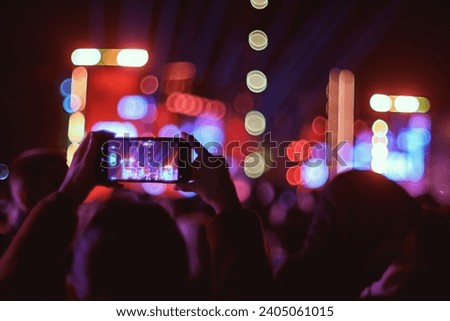 A person records a concert at Skanderberg Square at night with a smartphone, with bokeh stage lights in the background