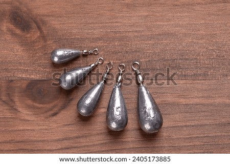 Fishing pear leads, also called sinkers, on a dark brown wood board, from above. Six different pear-shaped lead weights for fishing, from 5 to 20 grams, fused with wire and eyelets. From above. Photo. Royalty-Free Stock Photo #2405173885