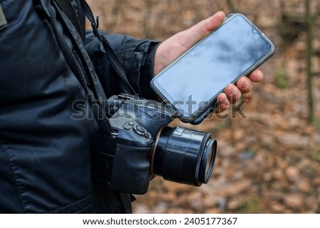 a man dressed in a black jacket with a black SLR old camera holds a new mobile phone in his hand on the street during the day