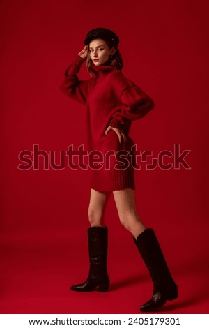 Fashionable confident woman wearing red winter knitted mini sweater dress, stylish cap, black knee high cowboy boots, posing on red background. Full-length studio fashion portrait