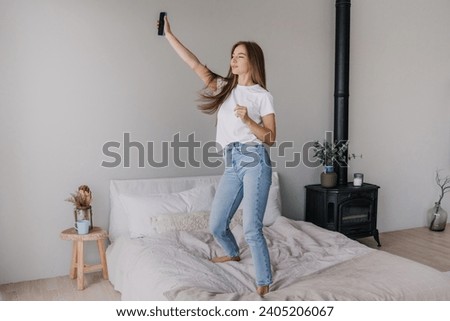 Joyful young woman in jeans and a white t-shirt taking a selfie on a bed, with a chic wood stove and plants in the background