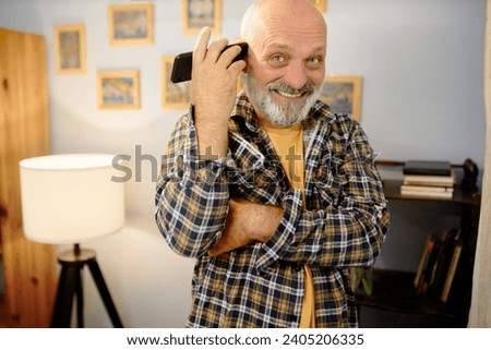 Happy smiling grandfather in plaid shirt listening to funny voice mail or audio message from his friends standing in his cabinet at home with folded arms, looking at camera with joyful face Royalty-Free Stock Photo #2405206335