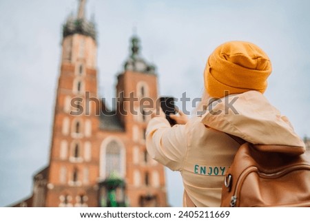 Back view of old senior female tourist is exploring new city. Blonde woman make a photo on Market Square in Krakow. Traveling Europe in autumn. St. Marys Basilica. High quality photo