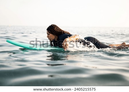 Profile picture of a dark-skinned young woman swimming on a surfboard in the sea dressed in a wetsuit. African women surfing. Water sports. Breaststroke swimming on a surfboard. Calm sea to practice.