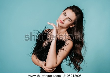 Sultry woman in a black feathered dress posing with her hand on her face, long wavy hair flowing, against a teal backdrop Royalty-Free Stock Photo #2405218005