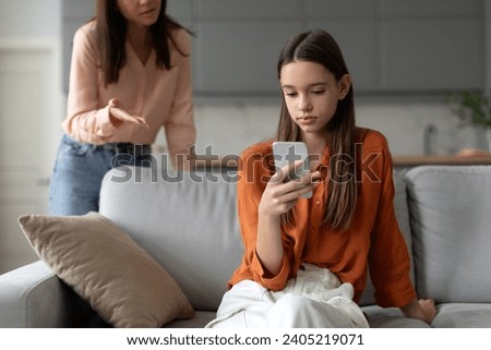 Angry mother shouting and arguing with teenage daughter over use of mobile phone, girl sitting on sofa at home, ignoring mom Royalty-Free Stock Photo #2405219071