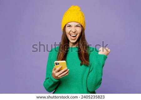 Young woman she wear green sweater yellow hat casual clothes hold in hand use mobile cell phone do winner gesture isolated on plain pastel light purple background studio portrait. Lifestyle concept