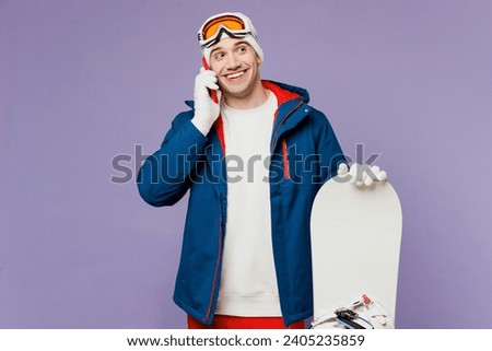Smiling man wear warm blue windbreaker jacket ski goggles mask hat talk speak on mobile cell phone hold snowboard spend extreme weekend winter season in mountains isolated on plain purple background