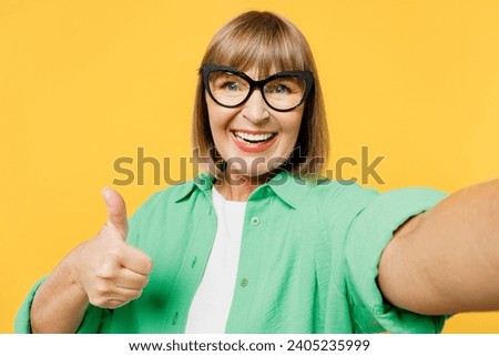 Close up elderly fun blonde woman 50s years old wear green shirt glasses casual clothes doing selfie shot pov on mobile cell phone show thumb up isolated on plain yellow background. Lifestyle concept