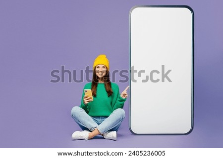 Full body young woman wearing green sweater yellow hat casual clothes sit point on big huge blank screen mobile cell phone with area using smartphone isolated on plain pastel light purple background
