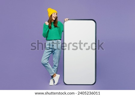 Full body young woman she wear green sweater yellow hat casual clothes big huge blank screen mobile cell phone smartphone with area do winner gesture isolated on plain pastel light purple background