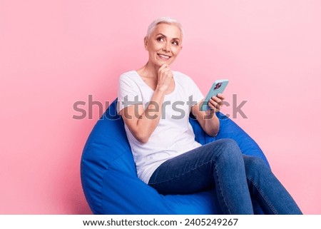 Full size photo of clever senior woman sit on bean bag hold smartphone look at offer empty space isolated on pink color background