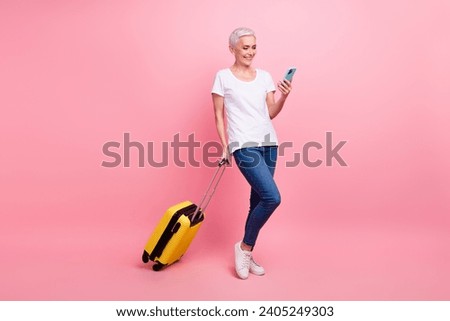 Full size photo of good mood woman wear white t-shirt jeans hold baggage booking tickets on smartphone isolated on pink color background