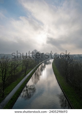 This image captures a tranquil morning scene over a river, taken from an elevated perspective. The soft sunlight diffuses through the hazy sky, reflecting gently on the water's surface. Flanking the