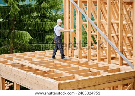 Carpenter constructing wooden-framed dwelling. Man measures distances with measuring tape while wearing work attire and headgear. The idea behind contemporary, eco-friendly building practices. Royalty-Free Stock Photo #2405324951
