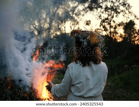 Woman in floral wreath near campfire in forest, dark nature background. rear view. Magic ceremonial, witchcraft. witch wiccan ritual for Beltane, Midsummer, Litha.