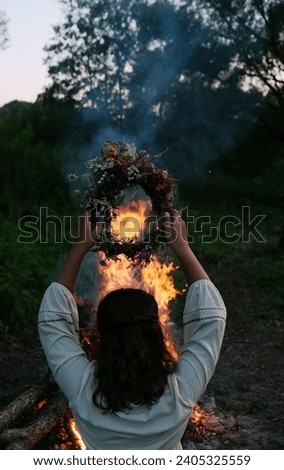 Girl with floral wreath in hands near campfire in forest, dark nature background. rear view. Magic ceremonial, witchcraft. witch wiccan ritual for Beltane, Midsummer, Litha.