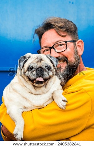 Portrait of man and dog. Young adult handsome with beard and eyewear hug and hold funny nice pug both looking on camera and blue background wall. Copy space and color. Love and friendship