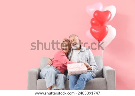 Mature couple with gift hugging on sofa against pink background. Valentine's Day celebration