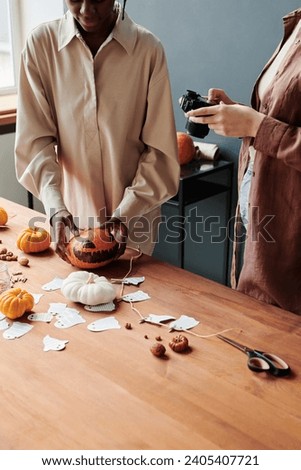 Young African American woman putting Halloween pumpkins on table next to other handmade decorations while her friend taking pictures