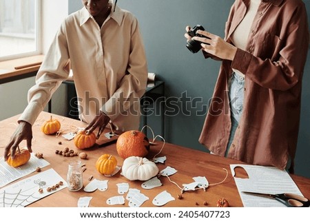 Young woman with photo camera taking shot of Halloween pumpkins, small funny paper ghosts, drawing of spider and other decorations
