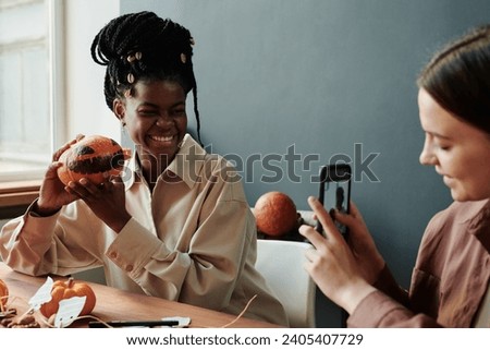 Happy young woman with Halloween pumpkin looking at smartphone camera while preparing for holiday and creating new decorations