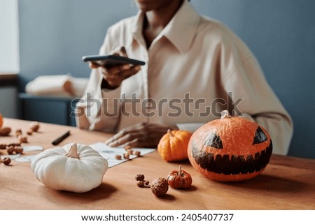 Spooky Halloween pumpkin with drawn scary face and other decorations lying on table against young woman taking picture of drawing