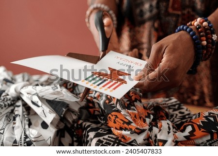 Hand of young unrecognizable woman cutting out new handmade Kwanzaa postcard over packed and wrapped giftboxes