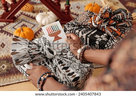 Hand of young African American woman holding creative Kwanzaa postcard with candleholder over packed and wrapped present