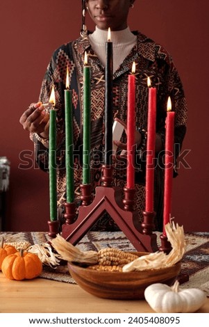 Young African American woman igniting seven multi-colored candles on candlestick standing on table with pumpkins and ears of corn