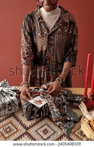 Happy young African American woman preparing Kwanzaa gifts and putting handmade postcard in wrap of giftbox at home