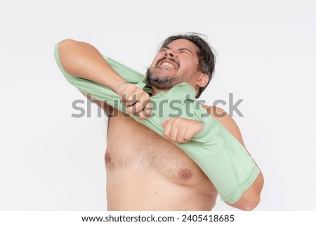 An overweight man struggles to put on an undersized polo shirt past his chest. A shirt too small or consequence of gaining too much weight. Isolated on a white background. Royalty-Free Stock Photo #2405418685