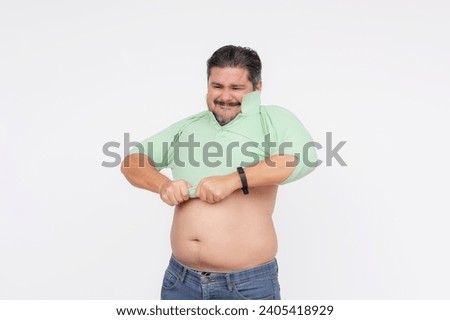 An overweight man struggles to put on an undersized polo shirt that used to fit him. A shirt too small or consequence of gaining too much weight. Isolated on a white background. Royalty-Free Stock Photo #2405418929