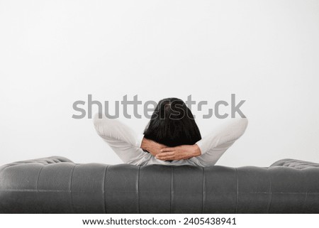 Rear view at relaxed woman with hands behind head sits on comfortable leather sofa on white wall background, feels mental balance, dreams of satisfaction, enjoys no stress free peaceful. Copy space Royalty-Free Stock Photo #2405438941