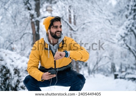 Confident handsome man with yellow beanie and windproof jacket tracking fitness progress on smart watch and using mobile phone while resting in squatting position after jogging. Royalty-Free Stock Photo #2405449405