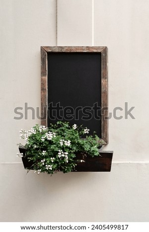 Empty dark wooden chalkboard, flowers in outdoor planter, hanging on wall facade of authentic retro cafe. Old European historical architecture building detail.