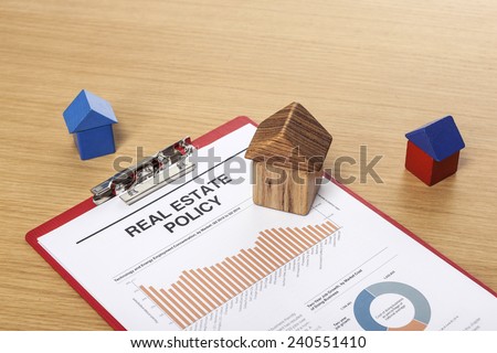 Three houses(made in wood block, red and blue) with graph paper(documents), clipboard on the office desk(table) behind white blind.