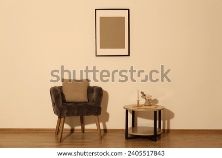 Comfortable armchair, cushion, table and decor elements in room with beige wall. Interior design