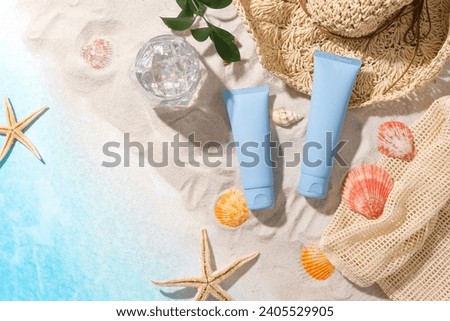Fresh sea background with white sand, starfish, seashells, water cup, straw hat, mesh basket and two striking blue cosmetic tubes. Seasonal cosmetic advertising.